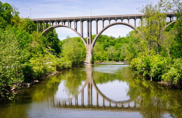 A large arched bridge spans over a calm river surrounded by green trees, with the bridge and trees reflected clearly in the water under a bright, partly cloudy sky.