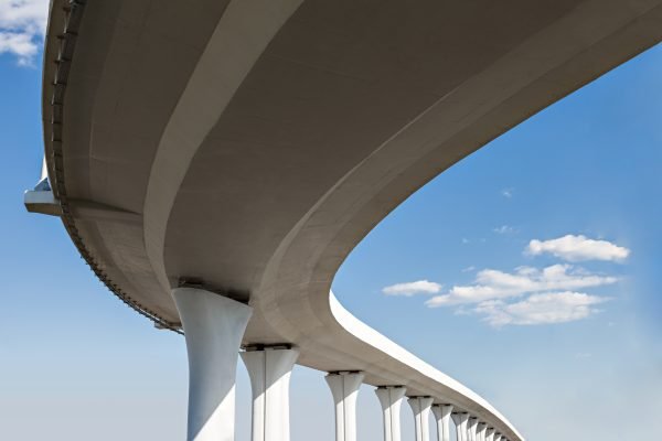A modern, elevated highway curves gracefully against a clear blue sky with a few scattered clouds, supported by tall white pillars.