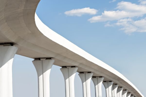 A modern elevated white bridge with smooth curves supported by tall columns, set against a blue sky with scattered clouds.
