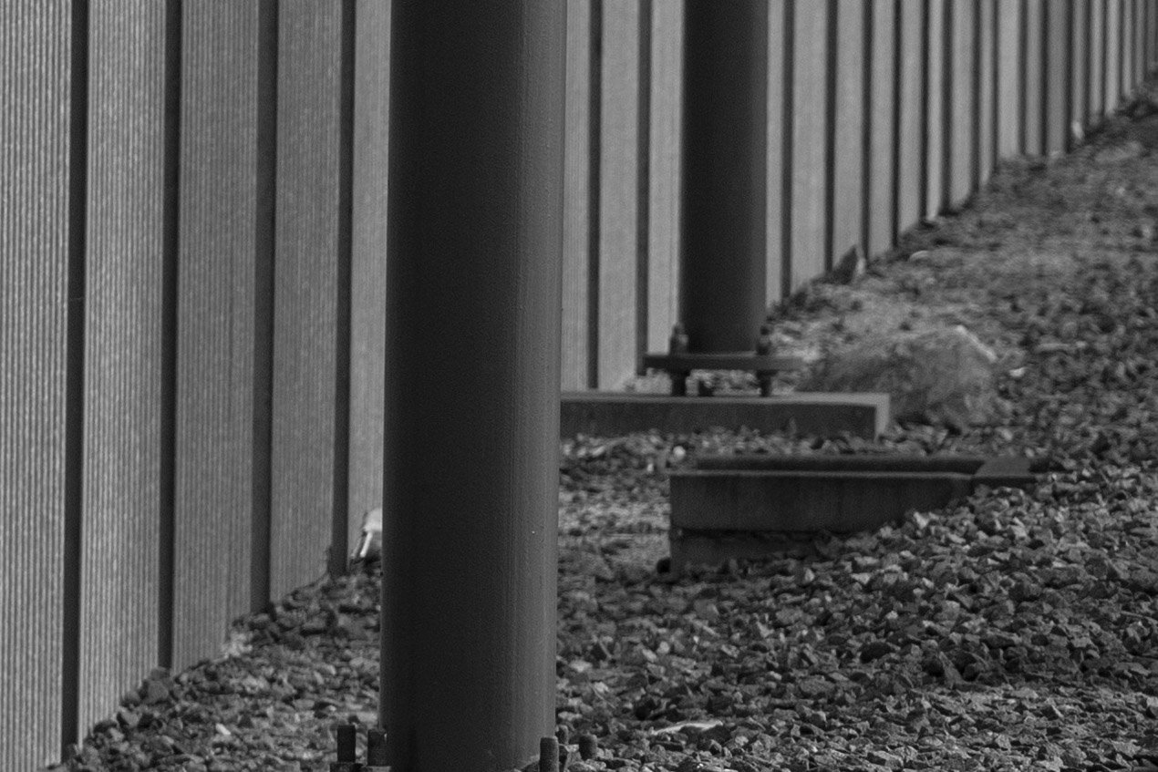 A black and white photo shows large metal poles supporting a tall, corrugated metal fence. The ground is covered with small rocks, and the poles extend into the distance, creating a repetitive pattern.