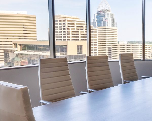 A modern conference room with four white chairs around a wooden table, large windows, and a cityscape with tall office buildings visible outside.