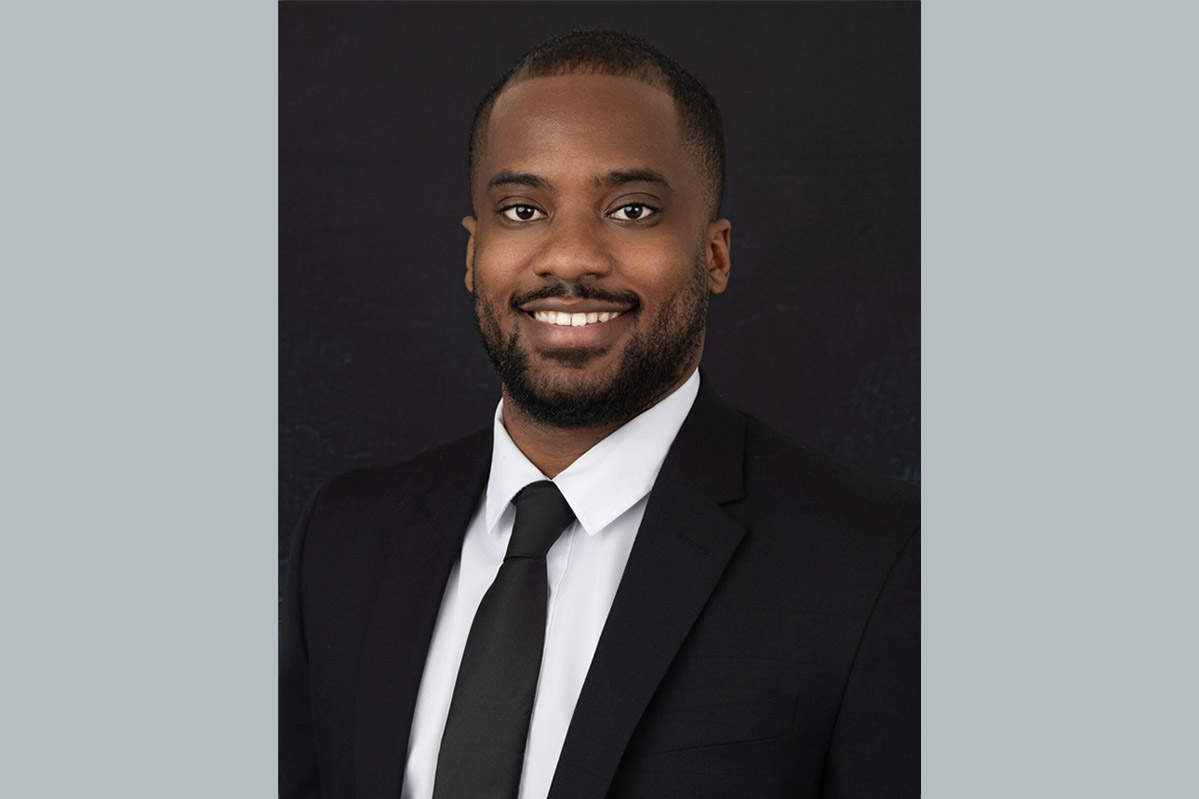 A man in a black suit, white shirt, and black tie smiles at the camera against a plain dark background.